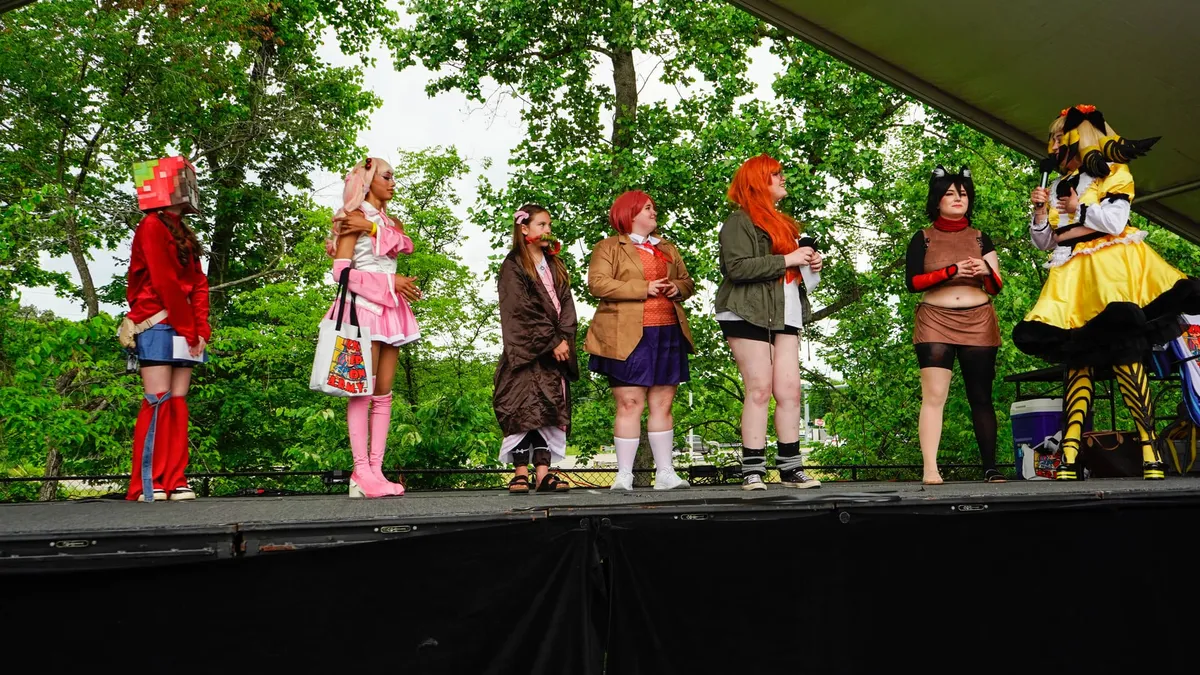 A lineup of about eight costumed cosplayers stands across an outdoor stage during a contest while a host in a yellow costume holds a microphone at one end at Knox Pop Con.